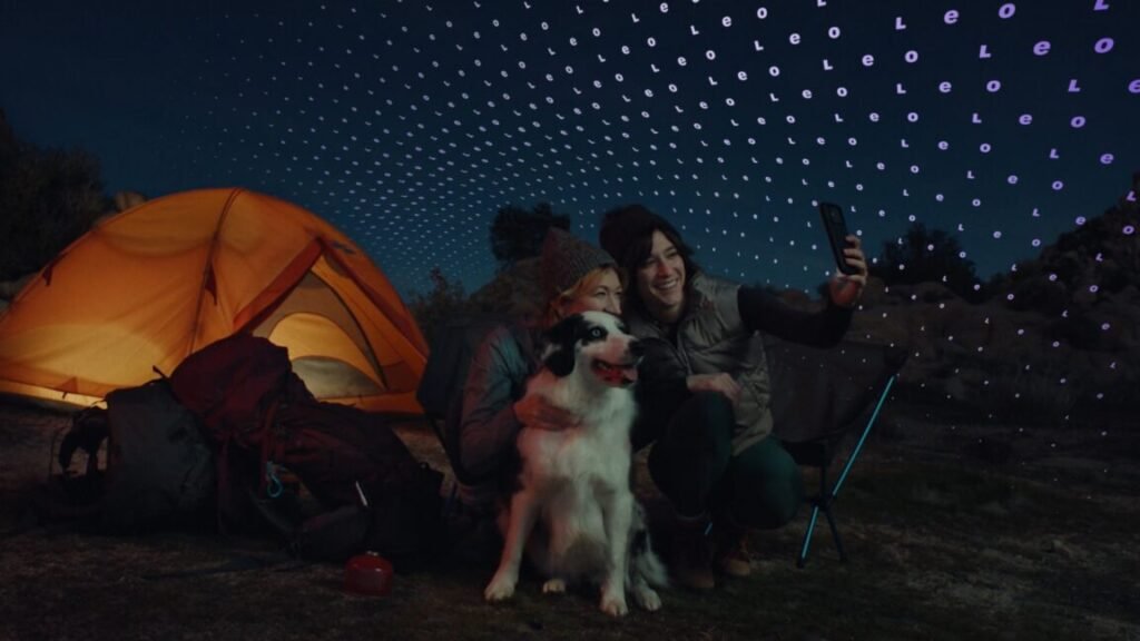 People camping at night take a selfie with a dog, using a phone connected to a satellite network.