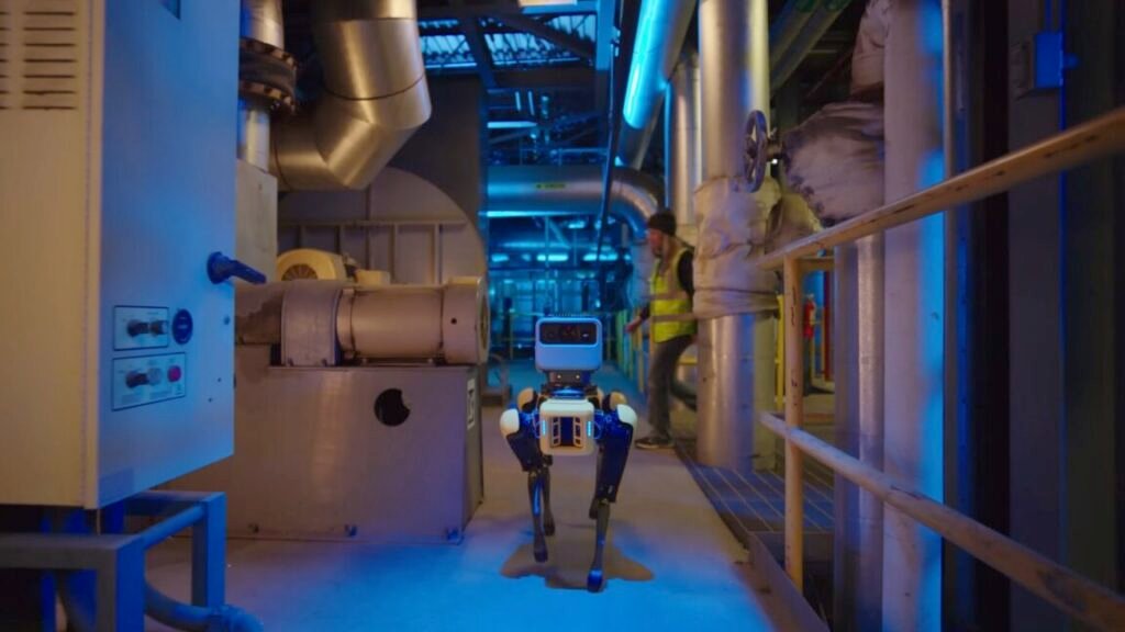 A four-legged Boston Dynamics robot called Spot walks down the hallway of an industrial facility. A human worker wearing a bright yellow safety vest is starting to cross the hallway behind the robot.