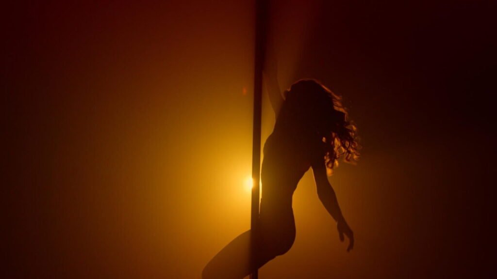 Flexible woman silhouette practicing poledance in nightclub spotlight. Sexual lady turning round pole making acrobatic tricks. Sensual girl pole dancing sexually in soft backlight