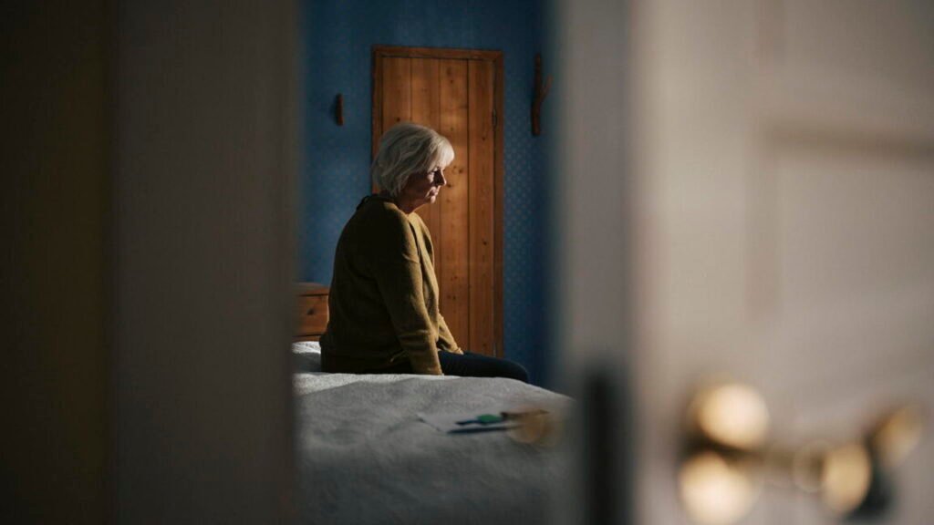 Sad elderly woman sitting on bed in bedroom at home