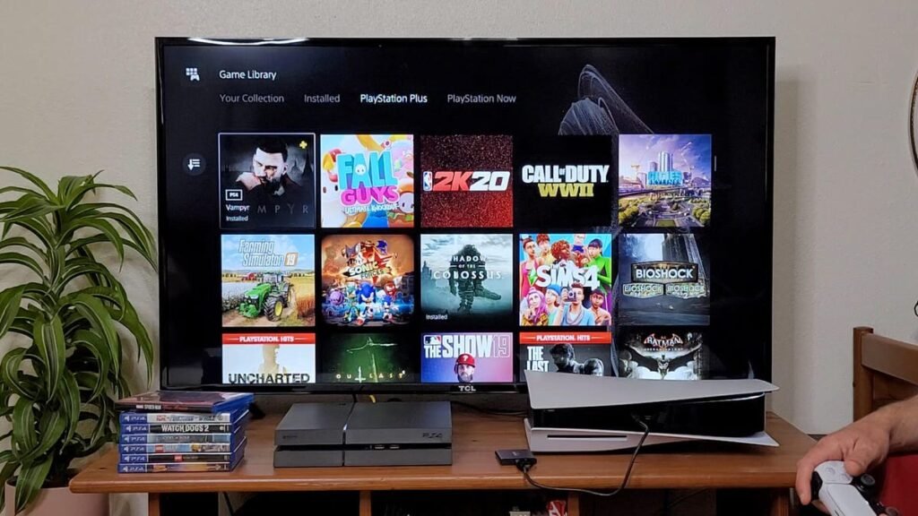 PS 4, PS 5 and game disc boxes on a table in front of a large TV screen showing PlayStation Plus selections