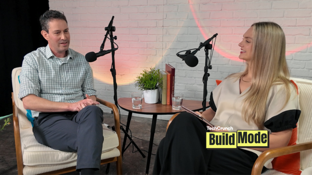 A podcast-style interview setup shows a man and a woman seated across from each other in armchairs, speaking into microphones. They sit beside a small round table with water glasses, books, and a potted plant, against a white brick wall backdrop. A “TechCrunch Build Mode” logo appears in the lower corner.