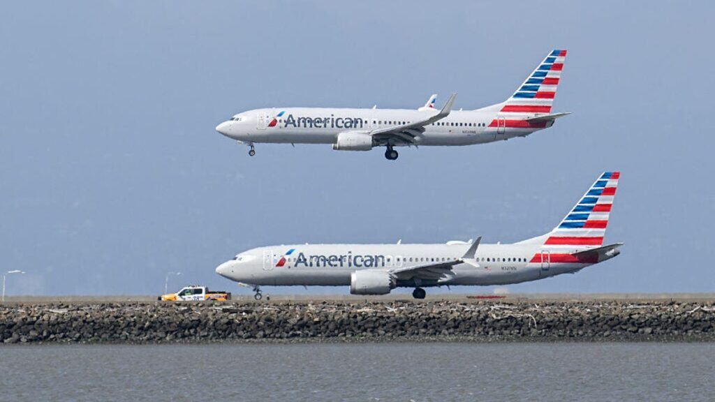 Two American Airlines planes are pictured at San Francisco International Airport. One is on the tarmac with a truck beside it. The other is currently taking off and gaining altitude.
