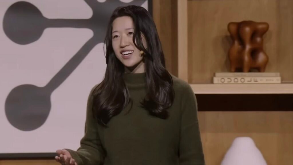 A woman with long black hair and a green sweater gestures and speaks in front of a wooden backdrop