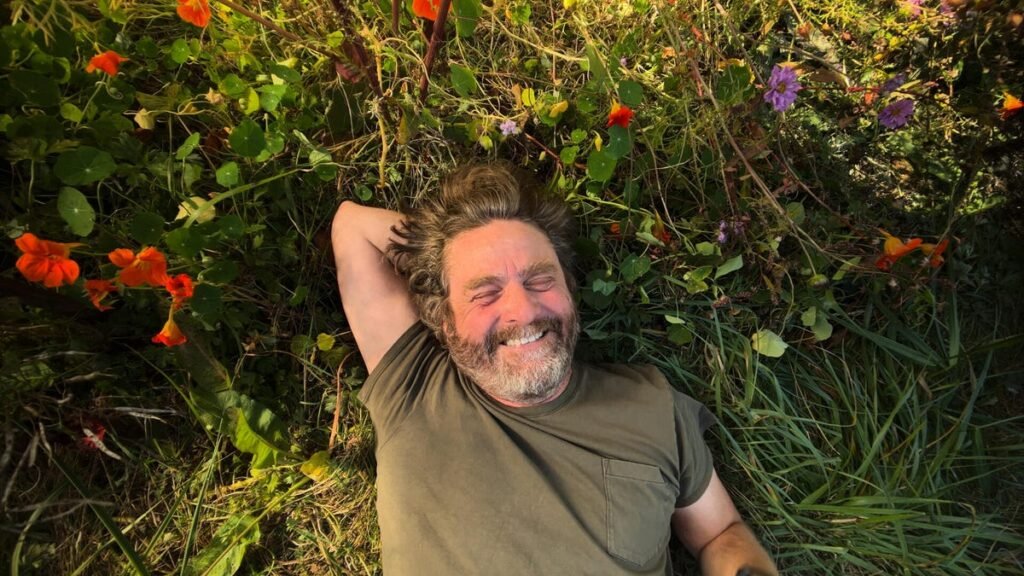 Zach Galifianakis smiles while lying in a field of flowers