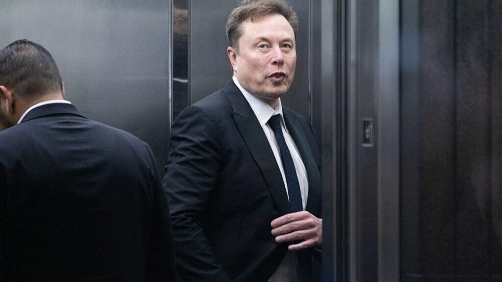 Elon Musk looks at the camera as he takes a deep breath, stepping out of an elevator. He is wearing a black suit and tie.