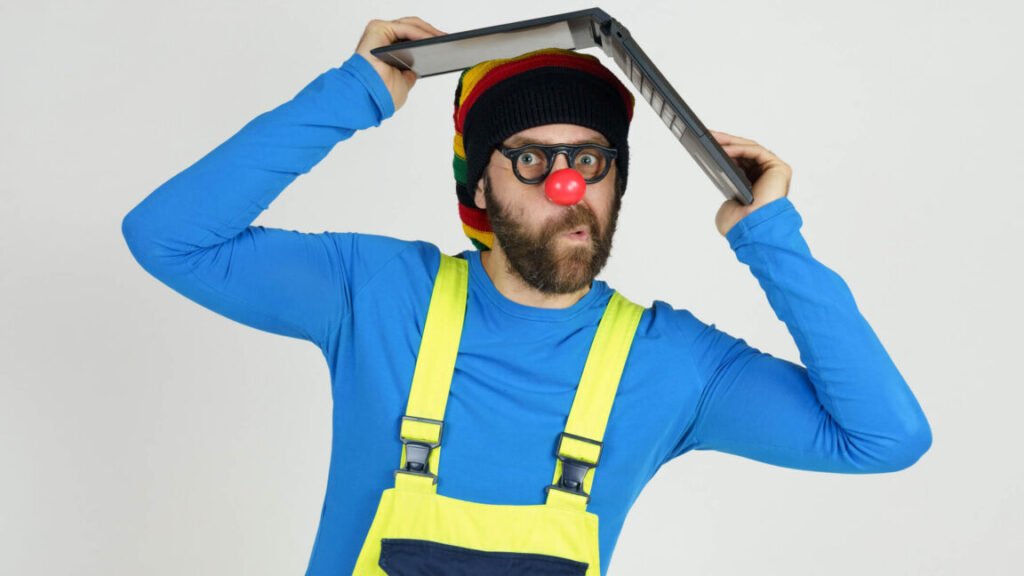 A clown in bright clothes holds a laptop above his head.