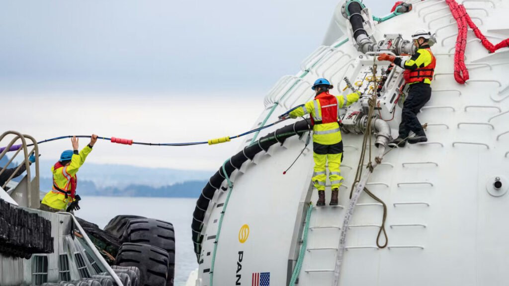 Two people stand on ladder rungs on a large, white sphere floating in the water as a third person standing on the dock or a nearby ship helps them handle a long cable. The white sphere is a prototype wave energy converter and floating AI "node" developed by the company Panthalassa.