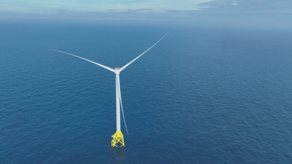 A white wind turbine with three blades each measuring 108 meters long stands against the backdrop of blue ocean waters stretching toward low-hanging clouds on the horizon.