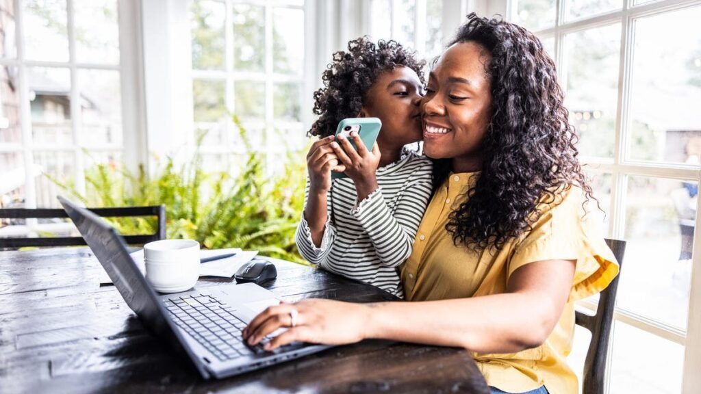 A woman smiles with a child while using a laptop on a black table.