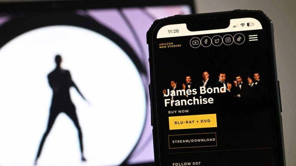 A phone screen shows an Amazon streaming page for the James Bond franchise. Behind it on a laptop screen is the iconic 007 gun barrel image.