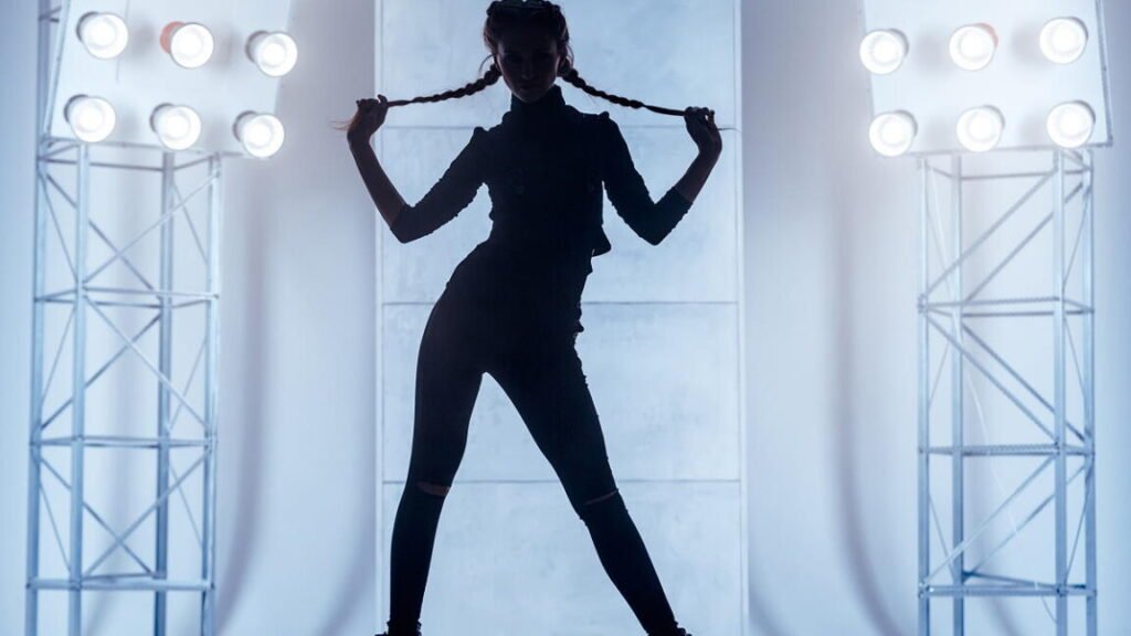 Beautiful slim young woman with two braids dressed in a black clinging clothes. The young woman dancer is performing the dance in the vogue style. She is standing holding the braids in hand. Studio shooting on a white background with light blue backlighting on a two stands