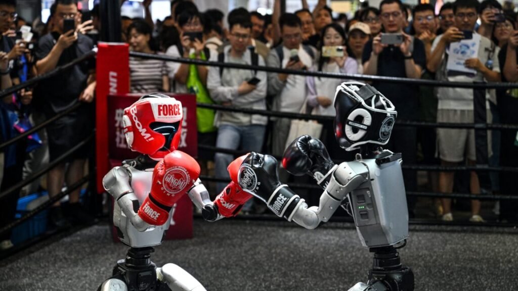 Humanoid robots controlled by a remote control by Unitree Robotics take part in a fight during the World Artificial Intelligence Conference (WAIC) in Shanghai on July 18, 2026. The foreground shows one robot on the left wearing a red helmet and boxing gloves and one robot on the right wearing black gear. The background shows a crowd of onlookers standing behind a boxing ring's ropes.
