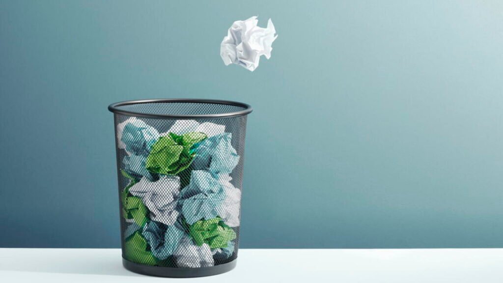A ball of crumpled paper flying towards a full waste paper basket