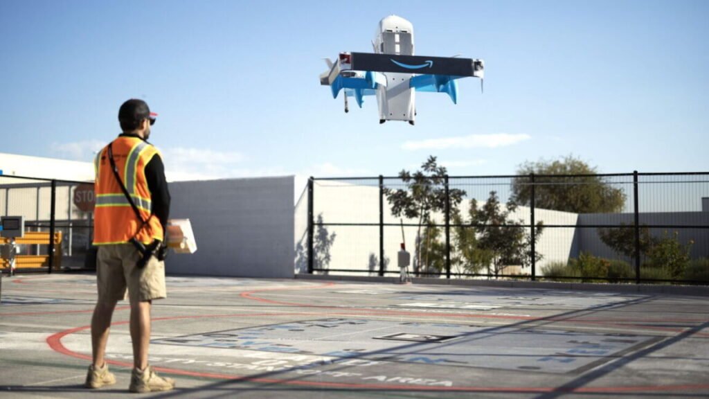 An Amazon Prime Air drone hovers above a paved lot in front of a person wearing an orange safety vest.