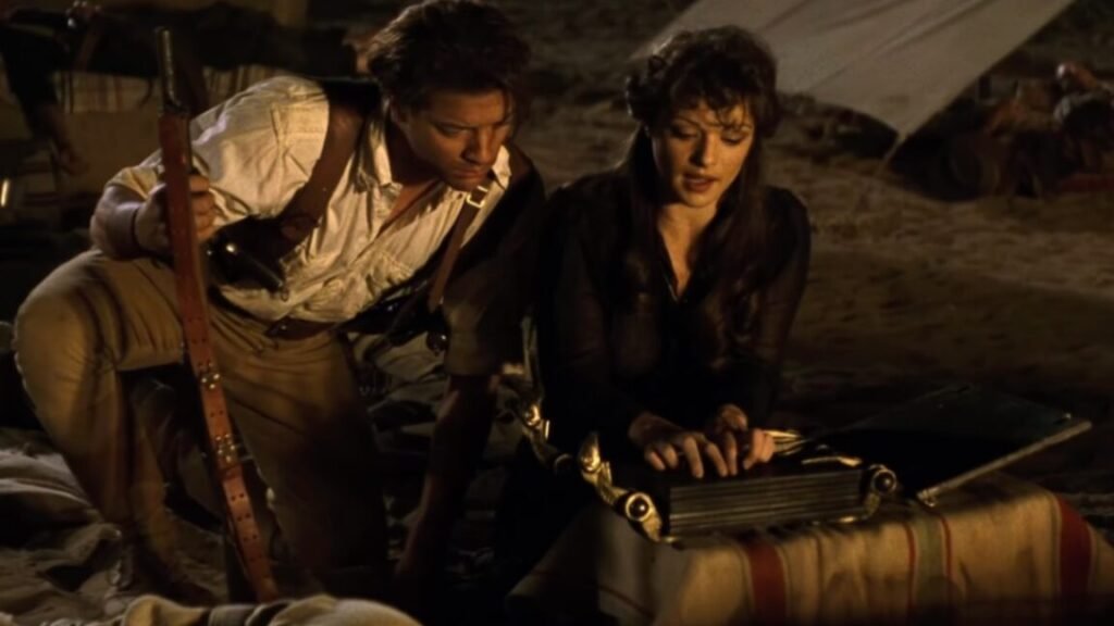 man in 1920s adventurer garb crouched near a woman beginning to read an ancient book at a desert camp site