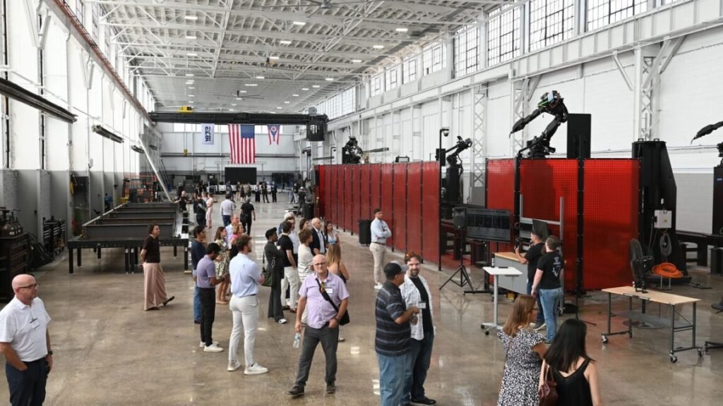 The inside view of a new factory opened by the startup 1872 on July 22, 2026. Welding workstations and robotic arms line the sides of the building while people mill around in the middle. A large American flag hangs against the back wall next to smaller flags representing the state of Ohio and the company 1872 logo.