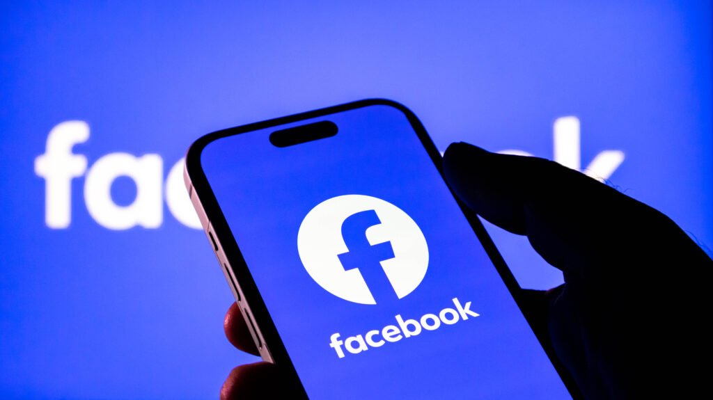 Closeup of a hand holding a smartphone displaying the facebook logo against a blurred blue background with the facebook brand name