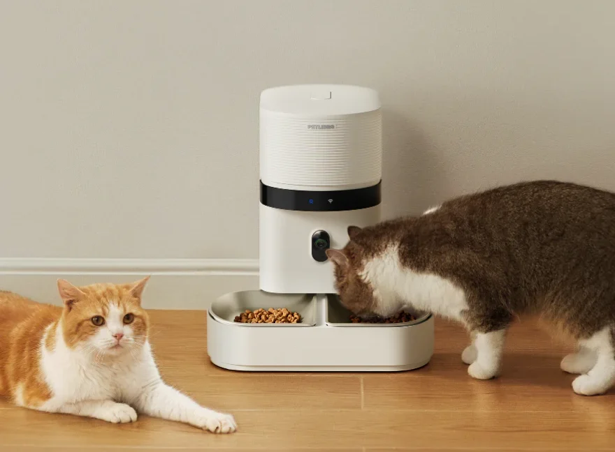 Petlibro accused of “gaslighting” users over smart pet feeder outage A cat sitting next to a Granary 2 that another cat is eating out of.