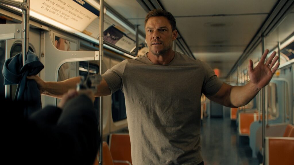 Alan Ritchson as Jack Reacher, holding up his hands on a train.