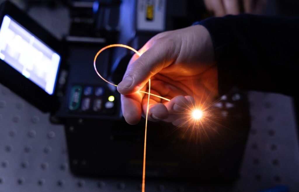A hand holds a single strand of fiber in a darkened room, with light pouring from its tip.