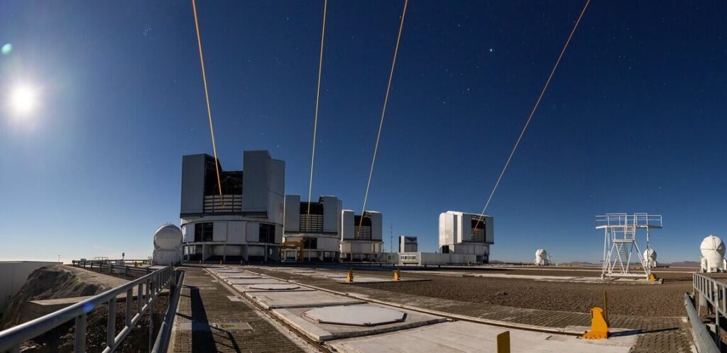 Image of four cylindrical buildings, each with an open slot for the telescope, and each emitting an orange colored laser