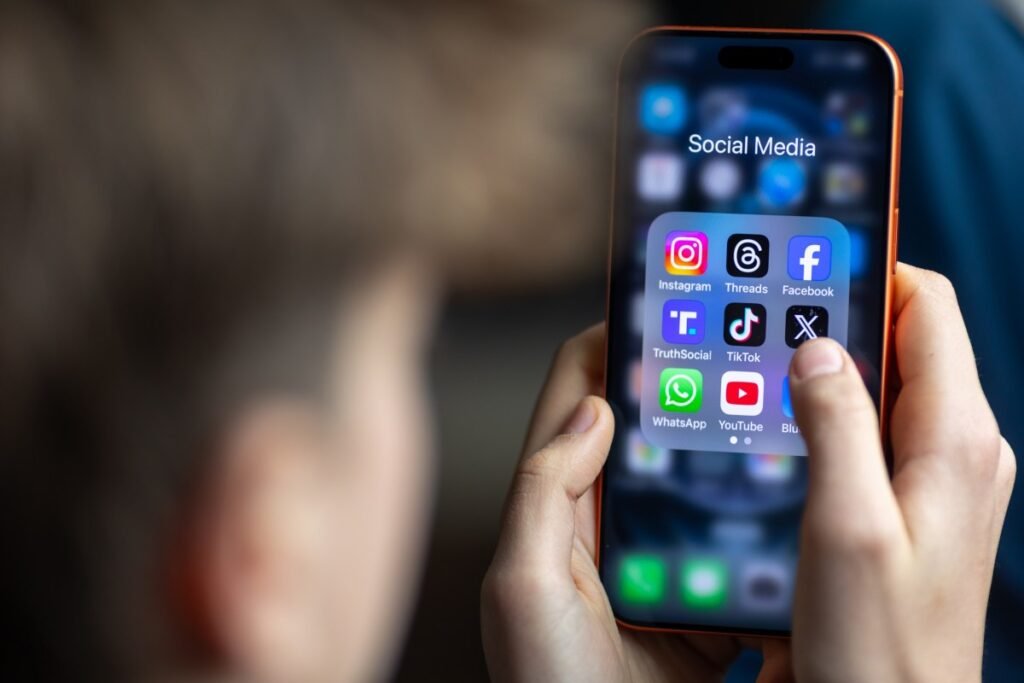a 14-year-old boy holds an iPhone screen displaying various social media and messaging apps
