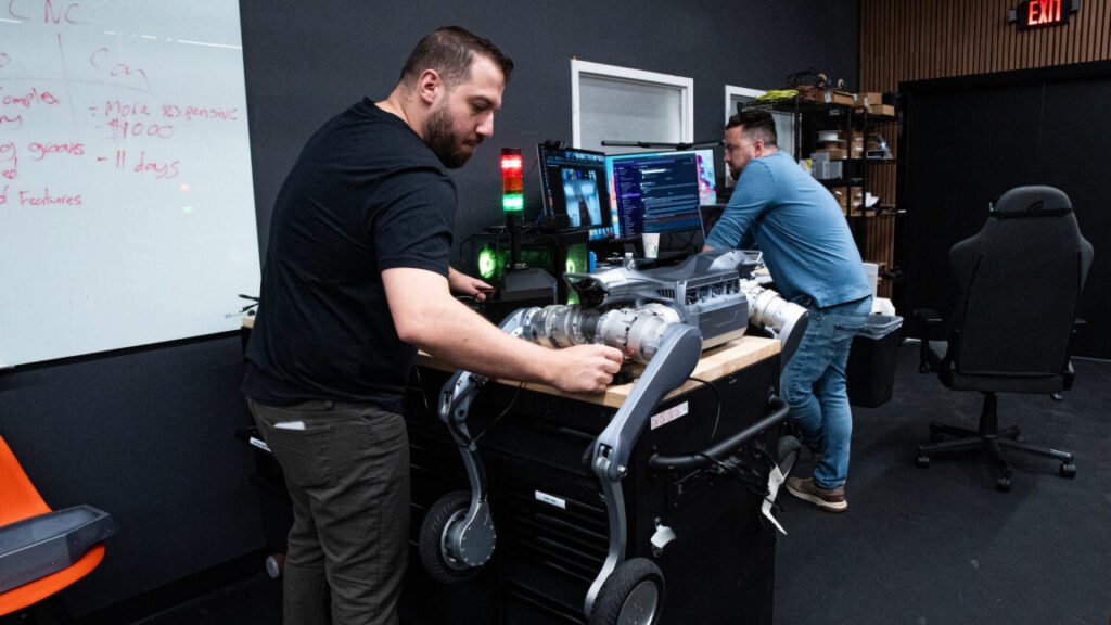 A person works on a quadruped robotic dog that is lying on a counter, while a second person stands in front of a computer with multiple display screens inside the office of Robo Inc.