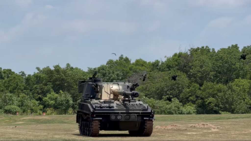 Quadcopter drones equipped with Auterion's Nemyx swarm autonomy system crash into a self-propelled gun sitting in a field with trees in the background during a test demonstration.