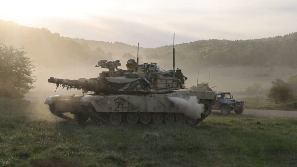 A US Army tank can be seen in profile with its main gun facing to the left. The background shows another US Army vehicle driving by with dust in the air. The scene took place during Exercise Combined Resolve held in Germany from April 9 to May 10, 2026.