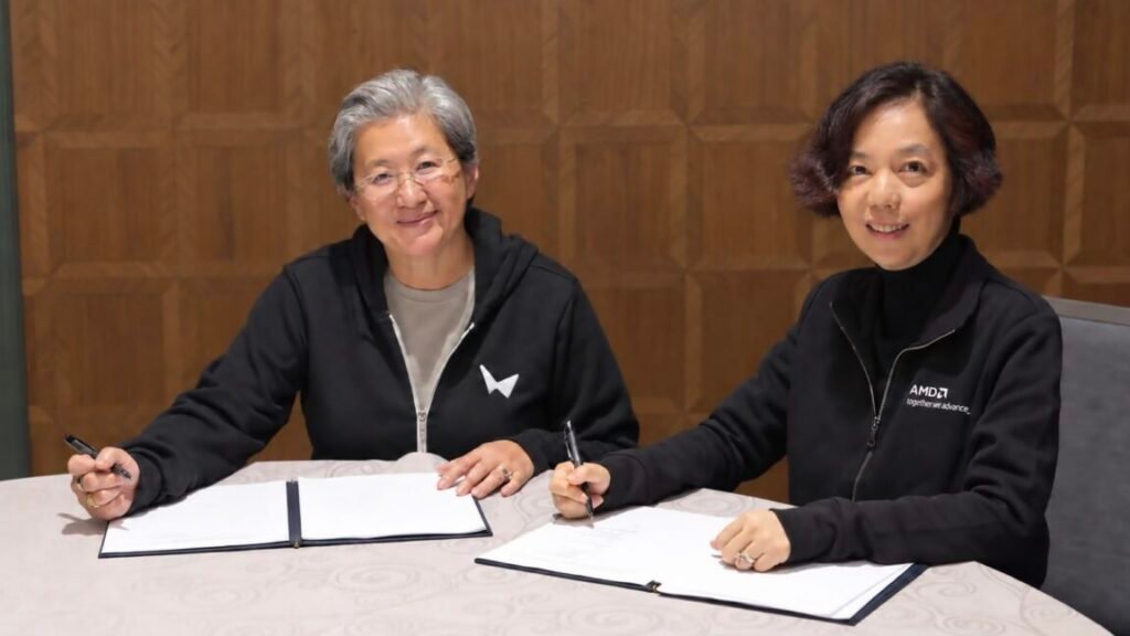 Two women in black hoodies—one from AMD wearing a World Labs hoodie, and one from World Labs wearing an AMD hoodie, smile at the camera while sitting a desk with papers to sign on it.