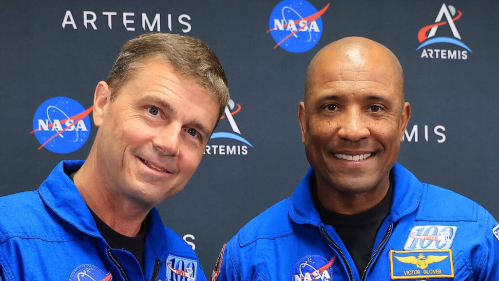 Two men in a blue flight suits pose for a photo in front of a black wall with printed logos.