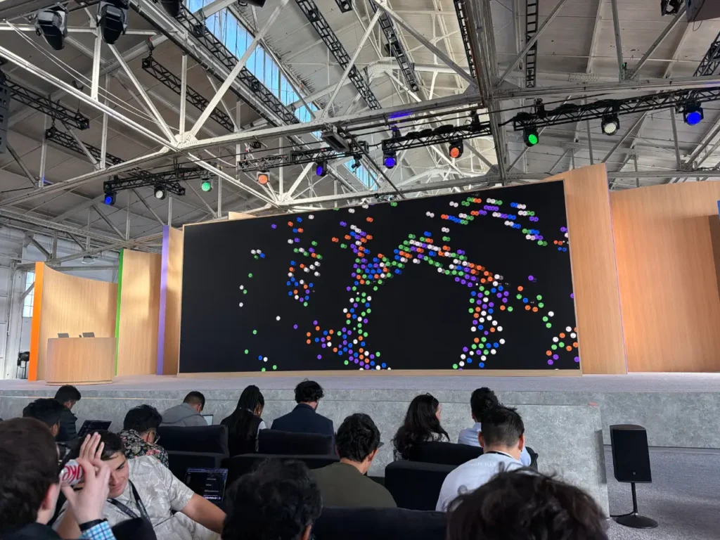 Attendees sit facing a large stage screen displaying a pattern made of colored dots in a bright industrial hall with exposed roof trusses and stage lights.
