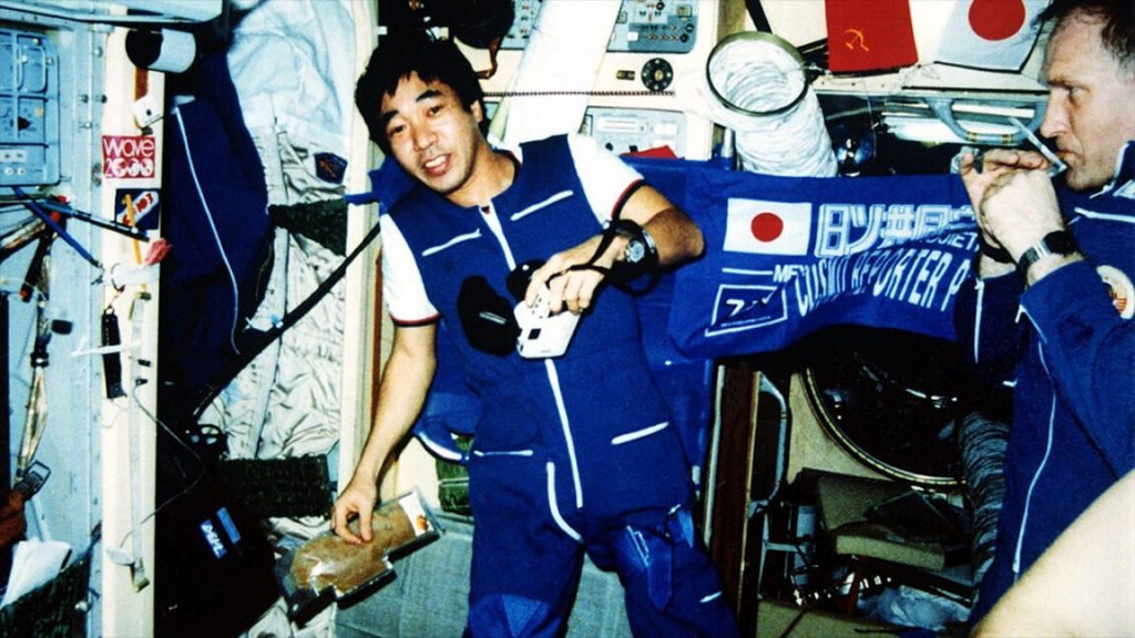 A man in a blue and white coveralls floats inside a space station module surrounded by instruments and stowage bags