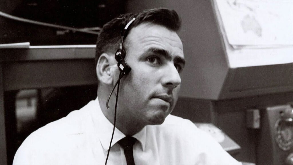 a black and white photo of a man in a white button-down shirt and thin black tie wearing a headset while seated in a room