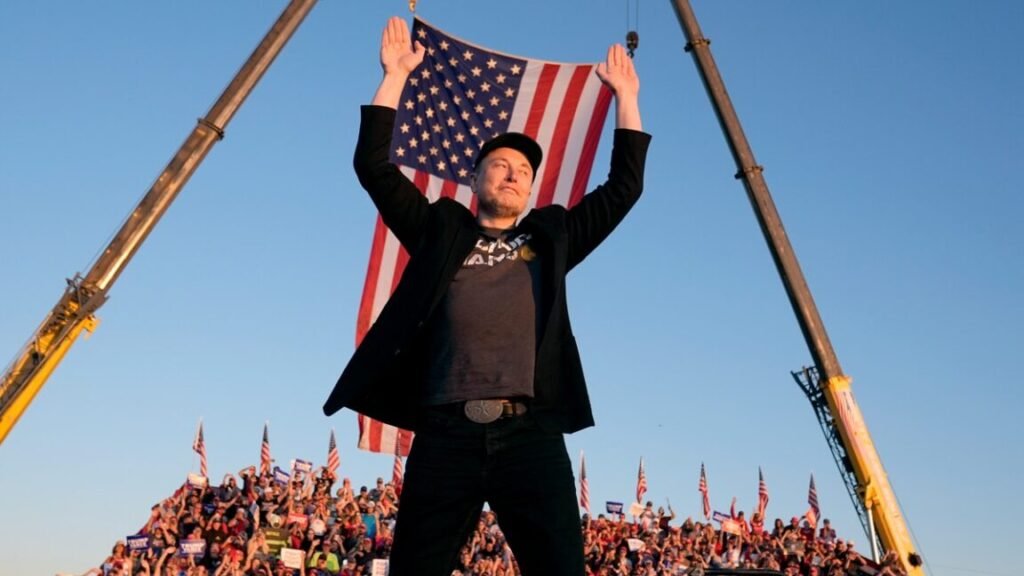 elon must in jeans and t shirt and denim jacket with a black MAGA hat in front of a US flag with arms raised as a crowd behind him cheers